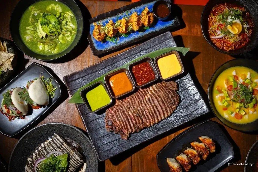 Overhead spread of Chinaloa dishes: aguachile, dumplings, bao and sushi on a dark table